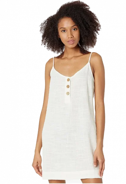 climate-friendly-beach-cover-up-rip-curl-dress@amazon A woman with curly hair is wearing a sleeveless, white summer dress with thin straps and three button details on the chest. Perfect for those seeking climate-friendly beach cover-ups, she stands confidently, looking towards the camera.