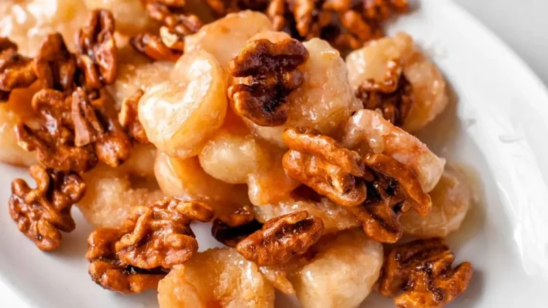A close-up of a plate of honey walnut shrimp. The shrimp are coated in a glistening light golden-brown sauce, and are mixed with glazed walnuts, all served on a white plate. The dish has a shiny, appetizing appearance.