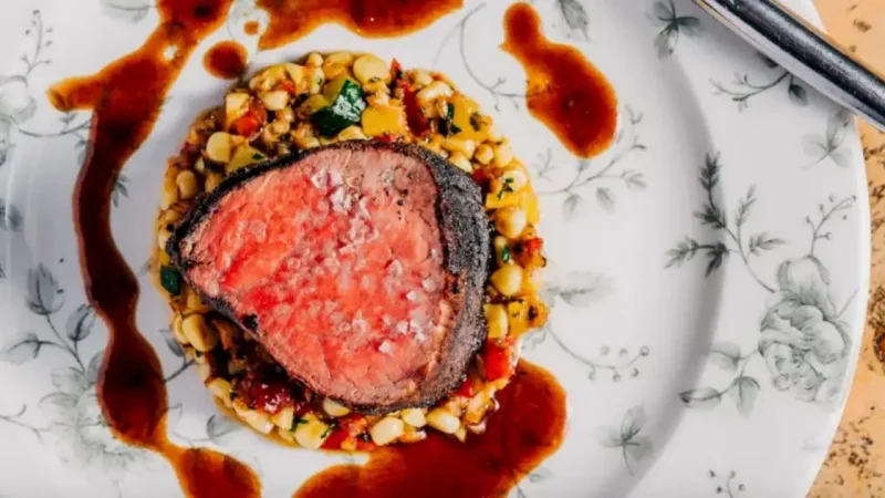 A succulent piece of medium-rare steak sits atop a bed of corn and vegetable medley, garnished with sea salt. The dish is artistically plated with a rich brown sauce drizzled around the edges on a white floral-patterned plate.