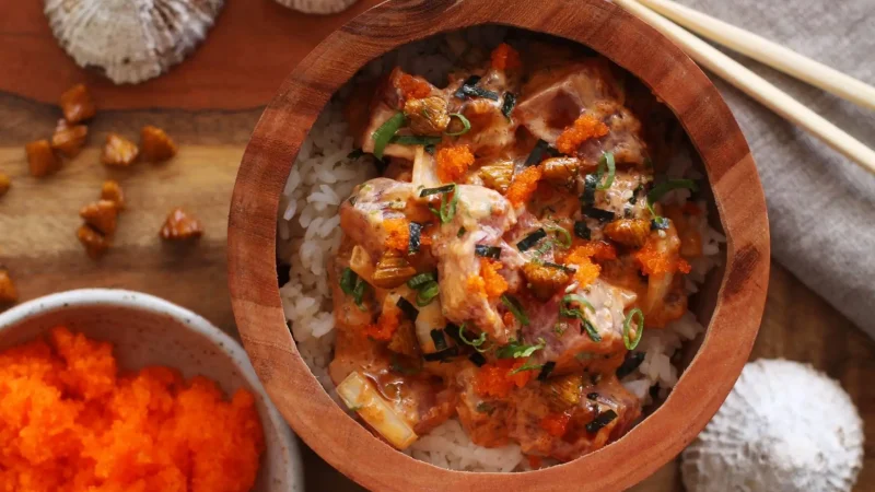A wooden bowl filled with a colorful dish of diced fish over rice, garnished with chopped green onions and bright orange roe. Surrounding the bowl are chopsticks, a small dish of roe, and decorative seashells. The setting exudes a rustic, coastal vibe.