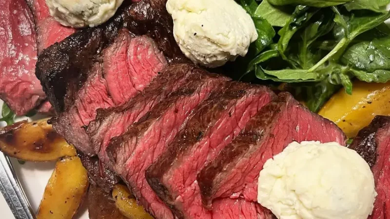 A plate of food featuring slices of medium-rare steak garnished with dollops of herb butter, roasted potato wedges, and a side of fresh green salad with arugula.