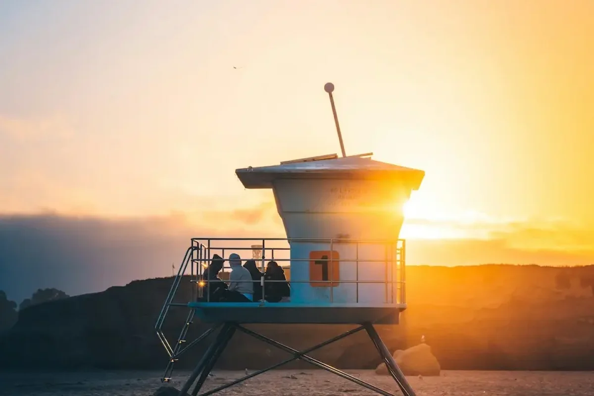A lifeguard tower on the beach is bathed in warm sunlight at sunset. Two people sit on the platform, facing the ocean along Central Coast California. The sky displays a gradient from deep blue to orange, with a hint of clouds in the background.