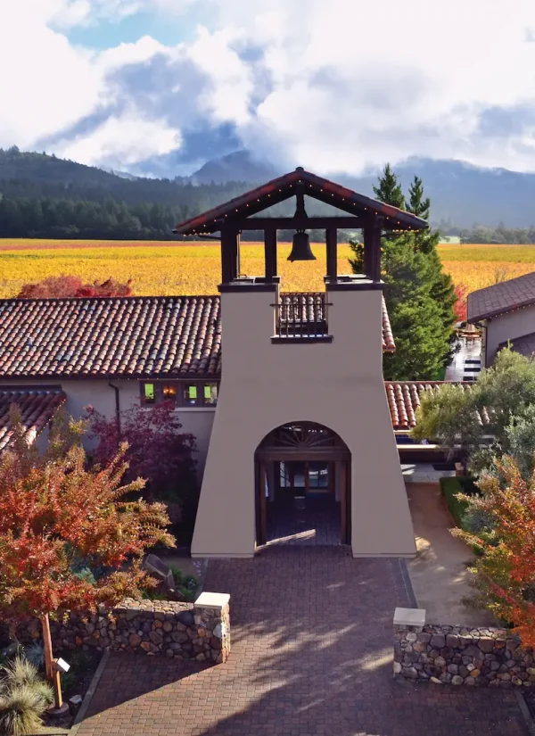 A vineyard estate with a tower featuring two bells stands amidst vibrant autumn foliage. Rows of grapevines stretch into the distance against a backdrop of rolling hills and a partly cloudy sky. The main building has a Mediterranean-style design with red-tiled roofs.
