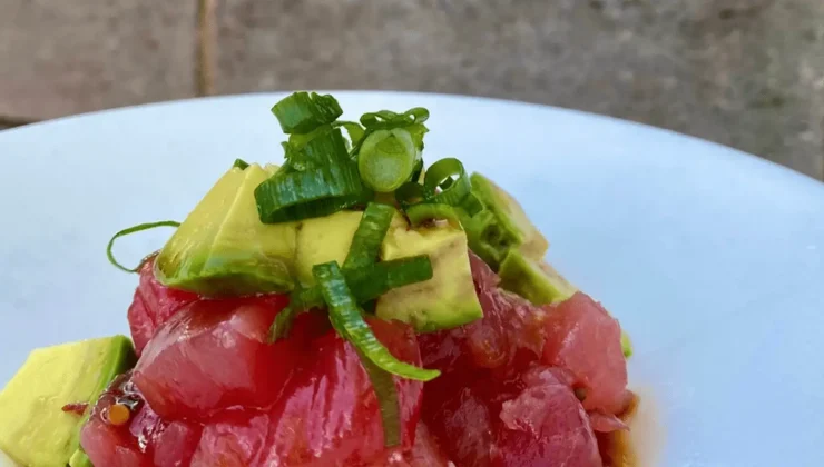 An up-close shot of a bowl of poke, featuring chunks of raw tuna, avocado, and green onions, all finely diced. The dish is placed against a neutral background with a stone or brick texture, highlighting the vibrant colors of the fresh ingredients.