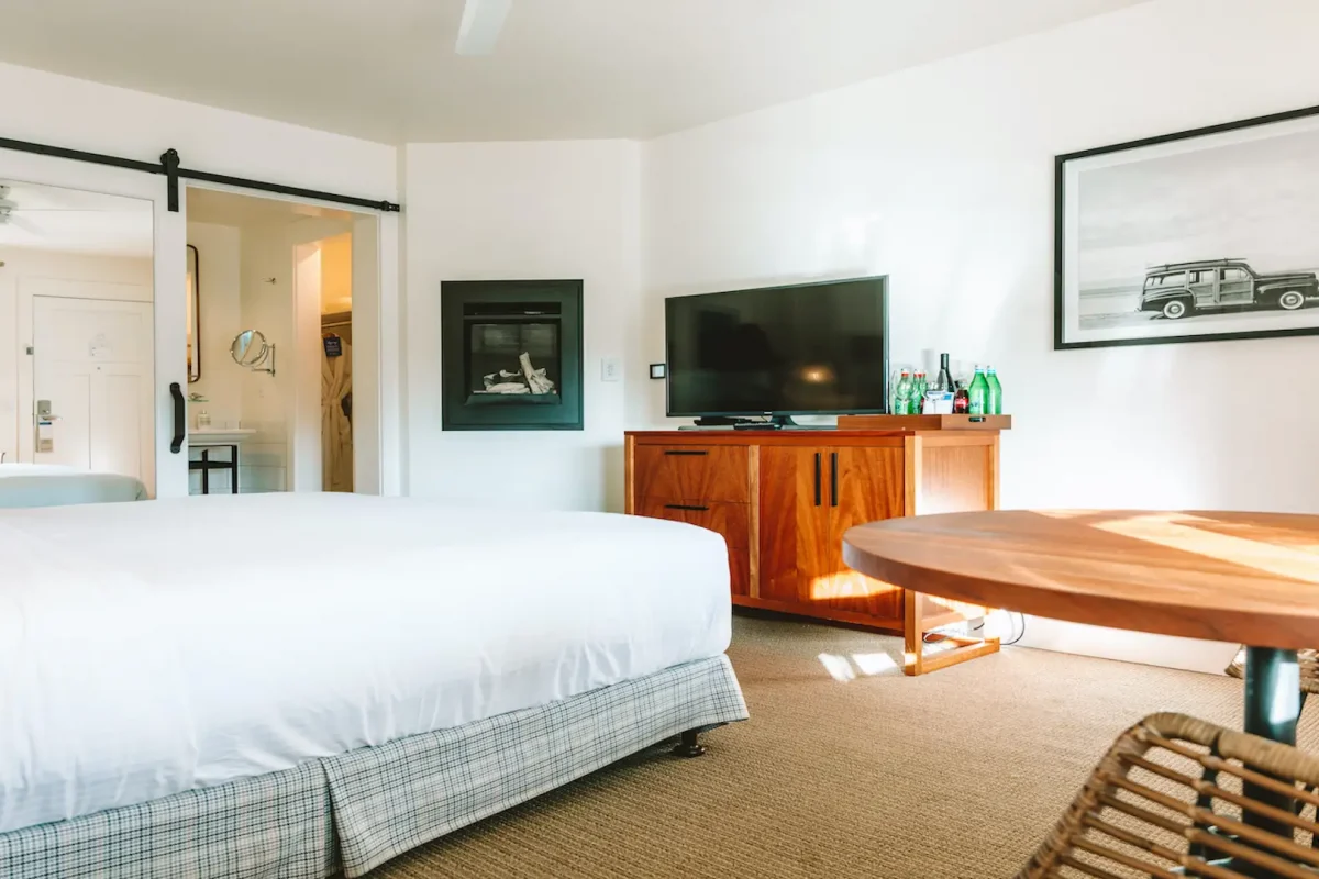 A bright and neatly arranged hotel room, one of the best value hotels in Carmel, featuring a large bed, flat-screen TV on a wooden stand, a round wooden table with a woven chair, and a wall-mounted photograph of a vehicle. The room also has sliding doors opening to a bathroom and visible amenities.
