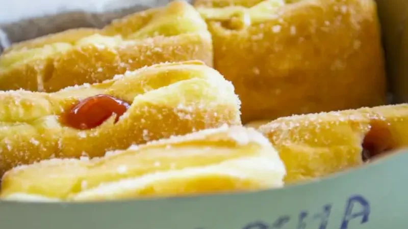 A close-up view of a box with the word "Aloha" printed on it, containing four sugar-coated pastries filled with a red filling, likely jelly or jam. One pastry is partially covered by a piece of parchment paper. They are placed on a green surface.